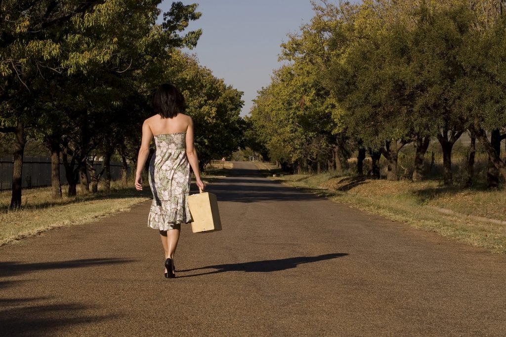 Brunette walking away from camera down tree lane with suitcase -freedom and wealth