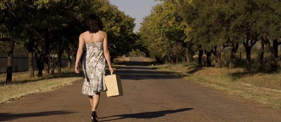 Brunette walking away from camera down tree lane with suitcase -freedom and wealth